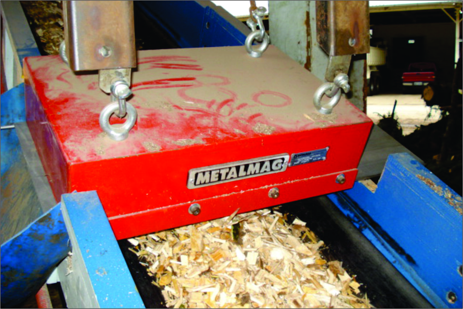 Separador magnético suspenso de limpeza manual instalado acima de uma correia transportadora, com estrutura metálica e correntes de sustentação em ambiente industrial.